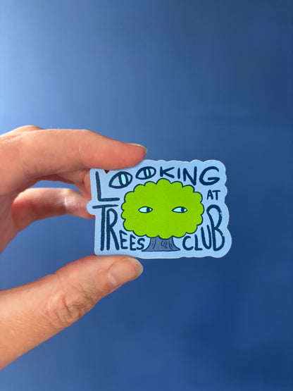 Hand holding a sticker with 'Looking at Trees Club' design against a blue background